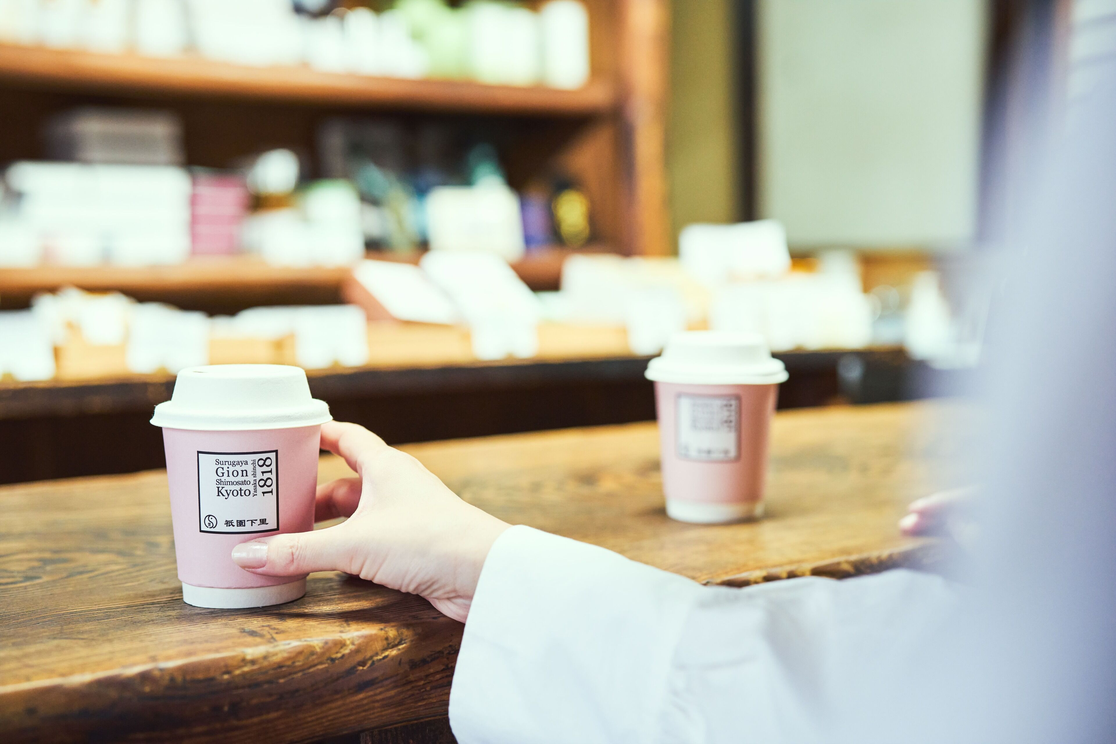 A hand holding a pink disposable coffee cup with a label, placed on a wooden table.
