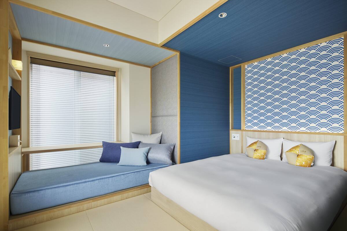 Une chambre d’hôtel comprenant un lit, un coin salon avec des coussins, une grande fenêtre avec des stores et un mur décoratif orné d’un motif ondulé.