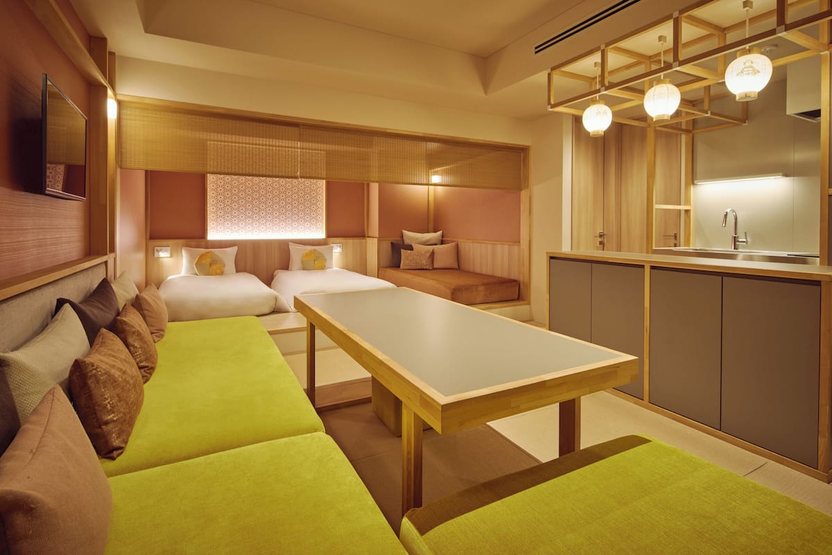 Une chambre d’hôtel moderne avec une tête de lit en bois, un grand lit, un canapé, une table à manger et un coin cuisine.