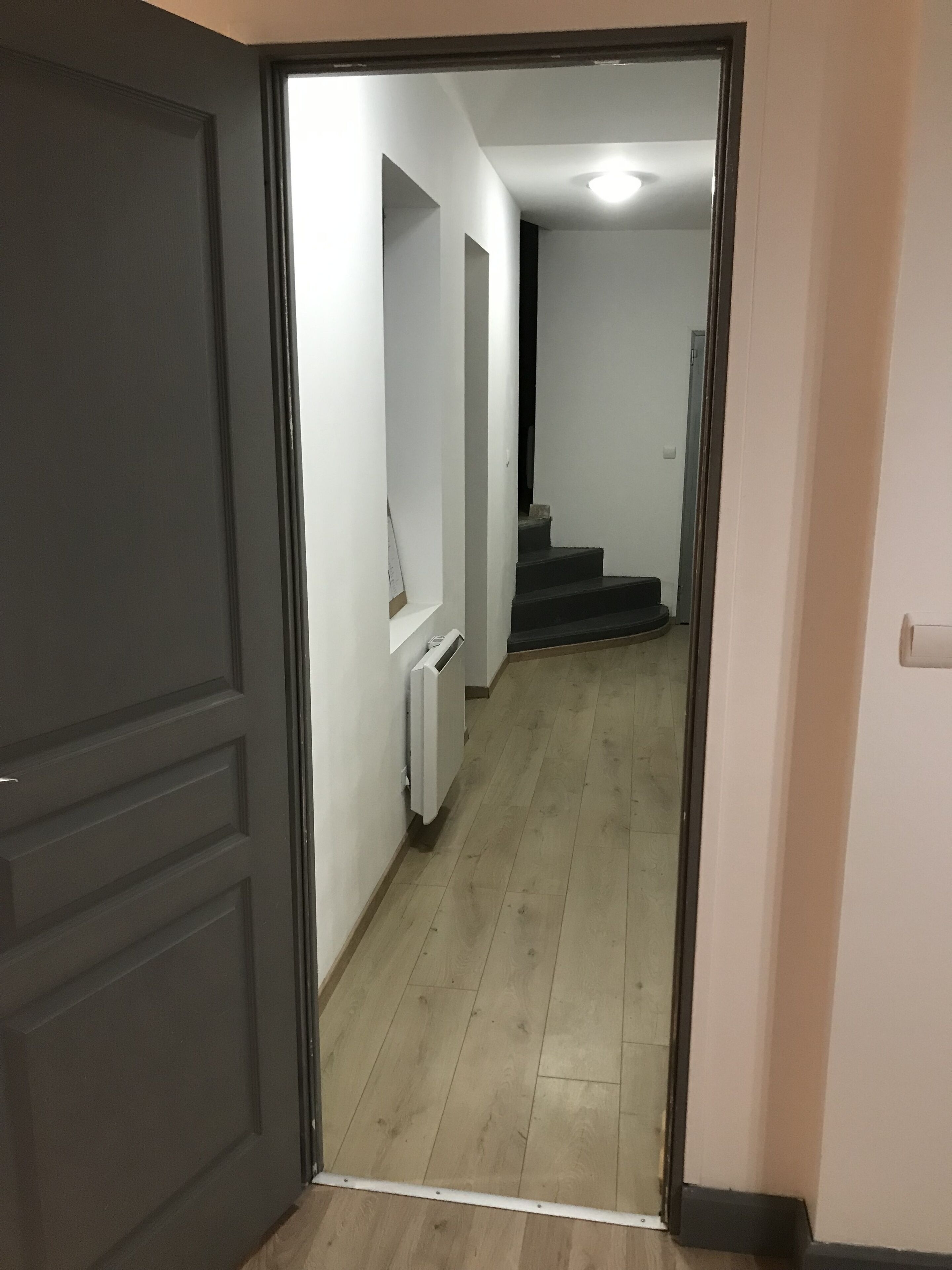 Un pasillo con una puerta oscura a la izquierda, un radiador en la pared y una escalera que lleva a otra habitación.