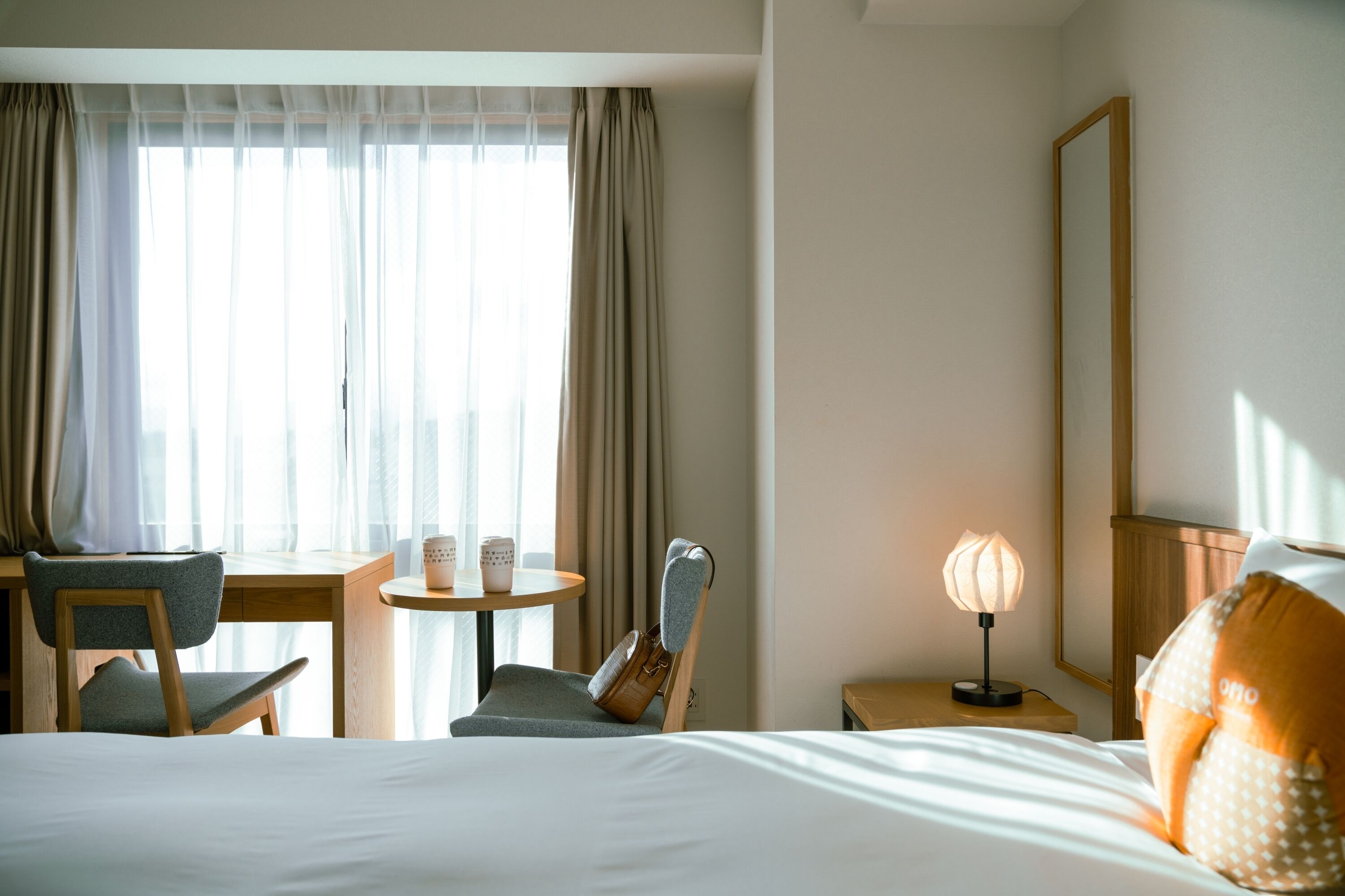 Una habitación de hotel con una cama, un escritorio con silla, una mesita de noche con lámpara y una ventana con cortinas.