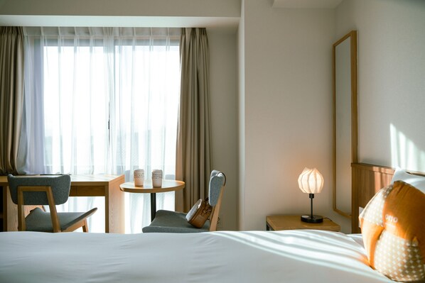 Une chambre d’hôtel comprenant un lit, un bureau avec une chaise, une petite table avec une lampe et une fenêtre avec des rideaux.