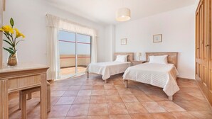 A hotel room with two beds, a desk, a vase with flowers, a window with curtains, and a ceiling light.