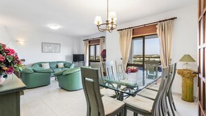 Una sala de estar con una mesa de comedor de vidrio, sofás verdes, un televisor y un balcón con cortinas.