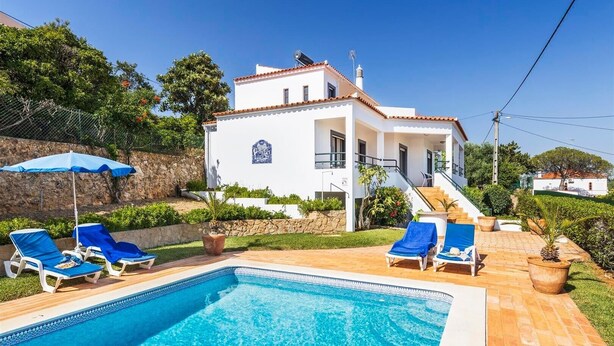 Una villa blanca con piscina, sillas de descanso azules y una escalera que lleva a la entrada.