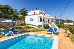 A white villa with a swimming pool, blue lounge chairs, and a staircase leading to the entrance.
