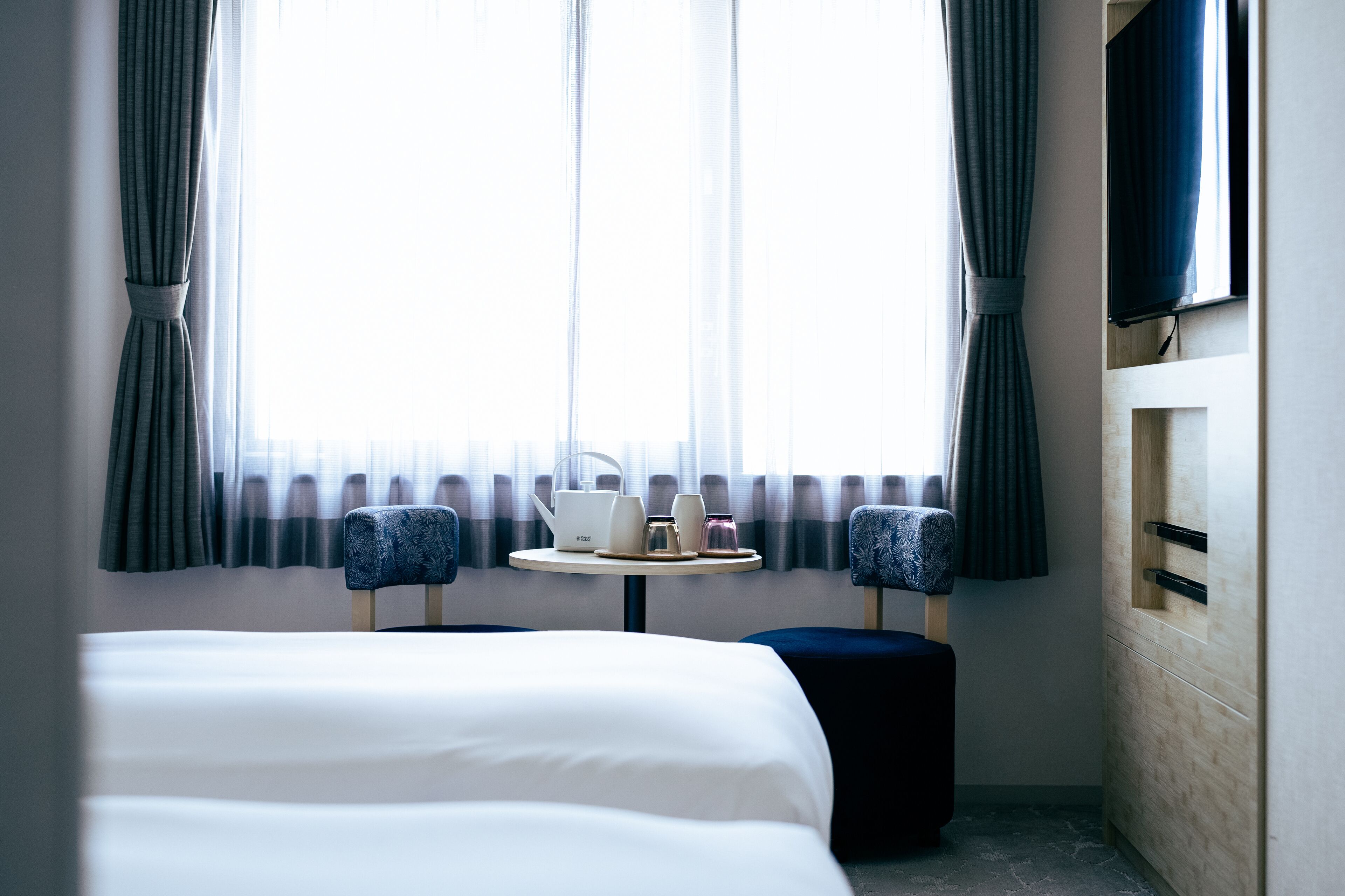 A hotel room with a bed, a small table with a kettle and cups, two armchairs, and a TV on the wall.