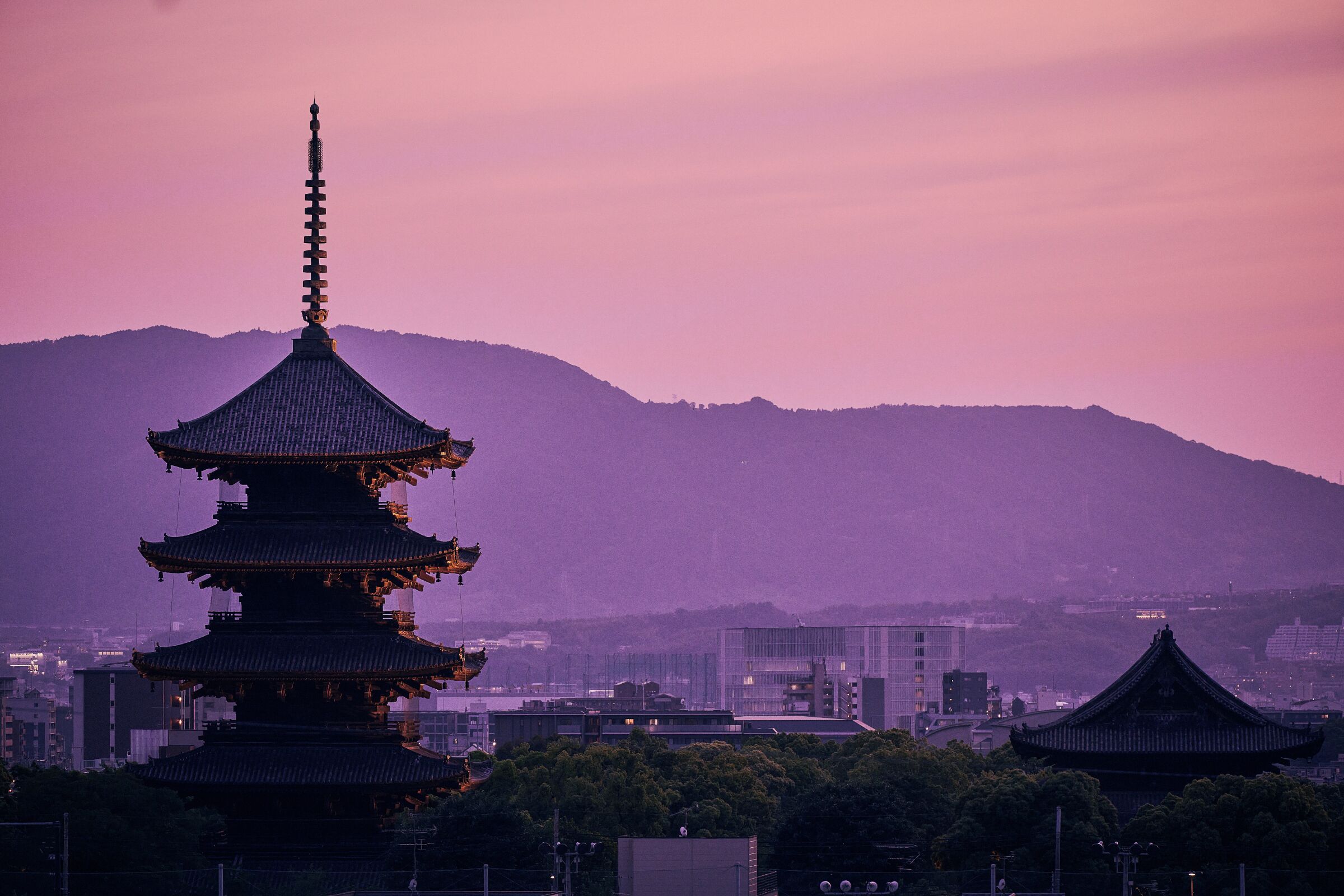 A traditional multi-tiered pagoda with a spire, set against a backdrop of mountains and urban buildings.