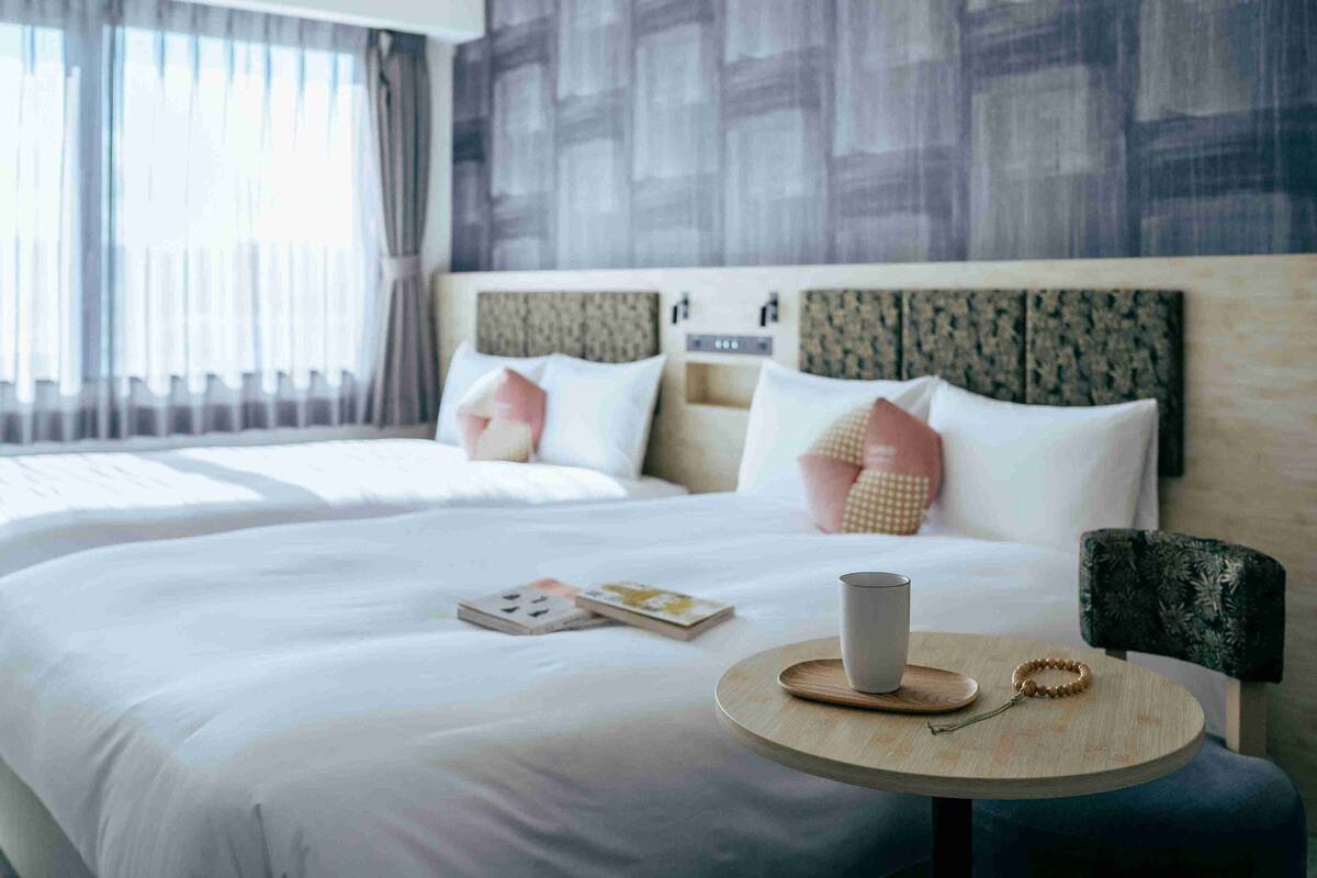 Ein Hotelzimmer mit einem großen Bett, einem Nachttisch mit Tasse und Buch sowie einem Sessel mit gemustertem Kissen.
