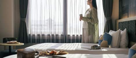 A hotel room with a bed, a small table with a cup, and a view of the outdoors through sheer curtains.