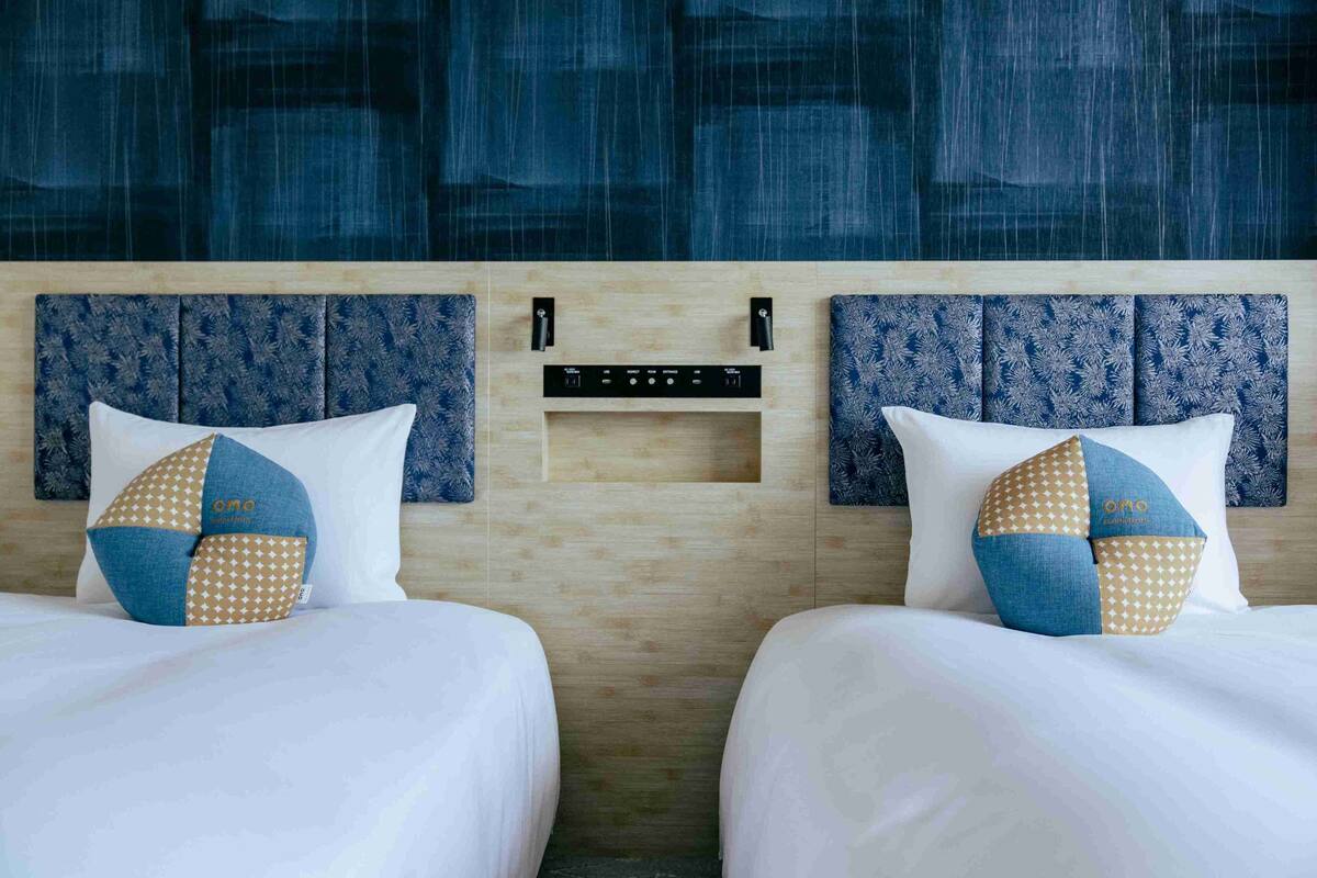 Ein Hotelzimmer mit zwei Betten, jedes mit einem gemusterten Kissen, einem hölzernen Kopfteil und einer Bedienkonsole an der Wand.