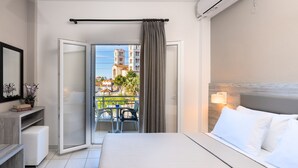 A hotel room with a bed, a desk, a chair, and a balcony with a view of buildings and palm trees.