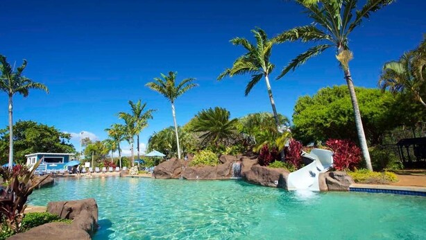 Una piscina cristalina rodeada de palmeras y rocas, con un área de descanso al fondo.