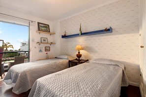 A room with two beds, a balcony, a table with a lamp, and wall-mounted shelves.