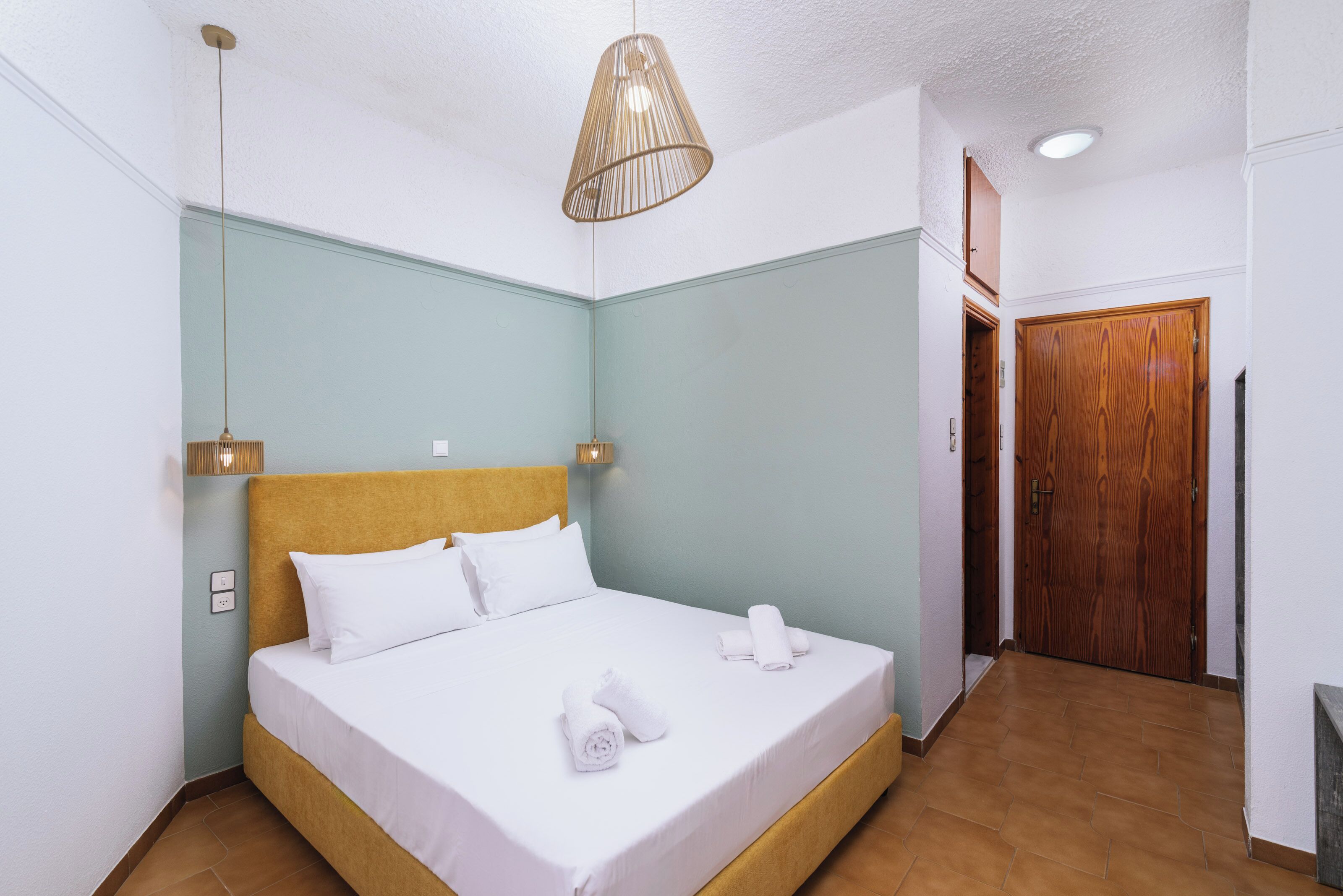 A neatly made bed with white linens and rolled towels, a wooden headboard, a hanging light fixture, and a wooden door in a room with light-colored walls and tiled flooring.
