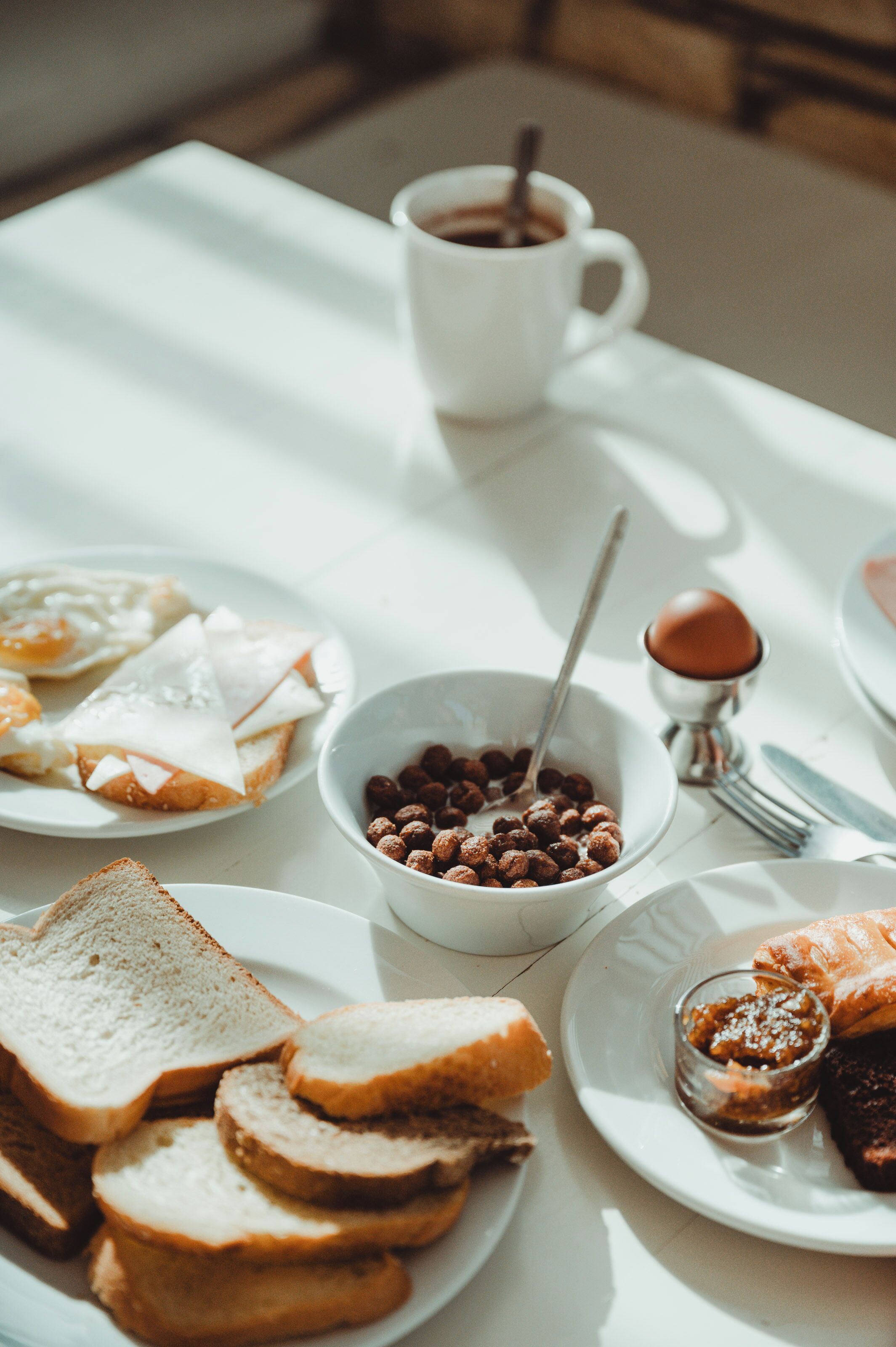 A breakfast table with toast, eggs, a bowl of cereal, a cup of coffee, and a small dish of jam.