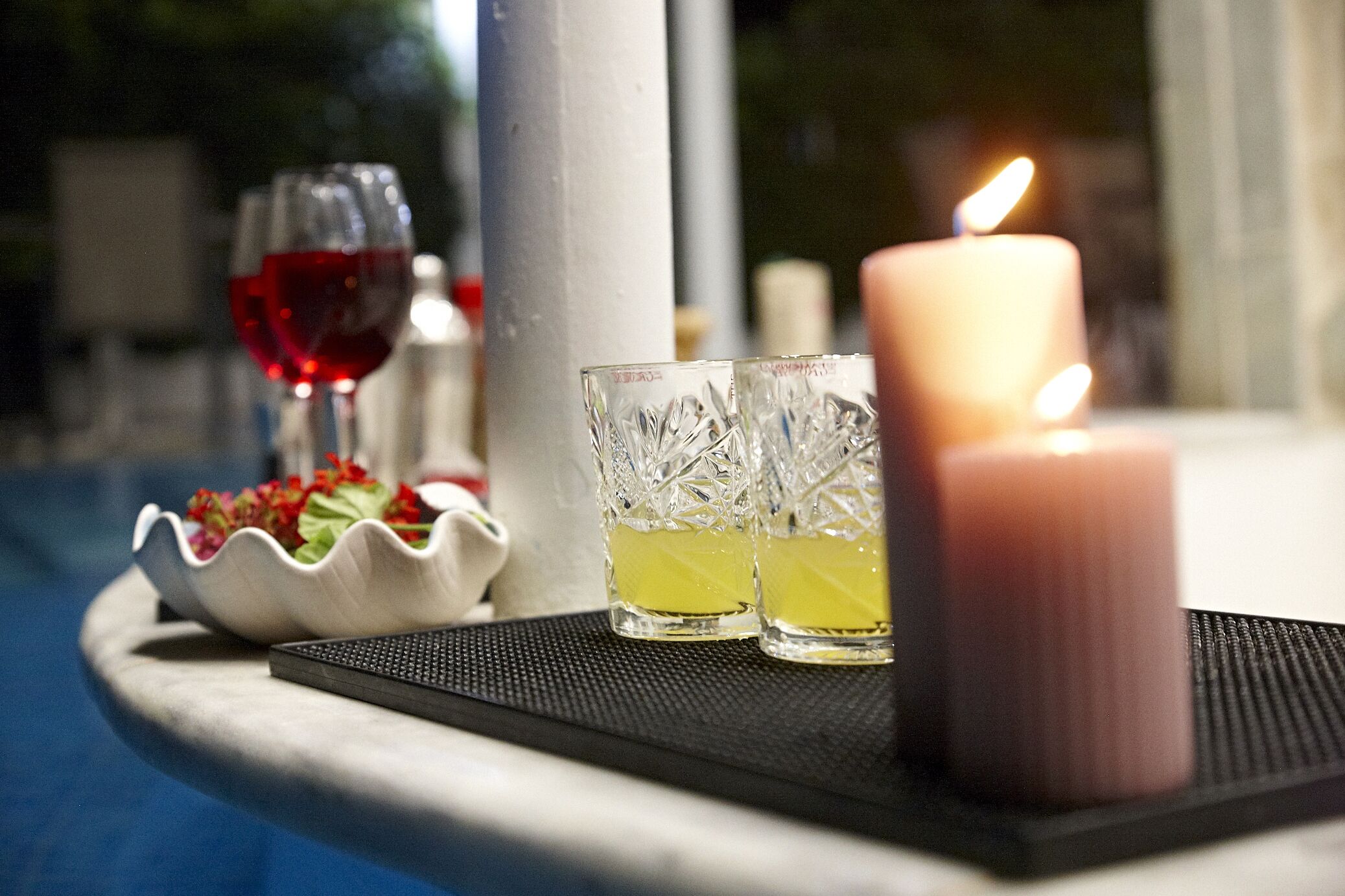 Et stearinlys, to glas og en skål med blomster på et bord i nærheden af en pool.