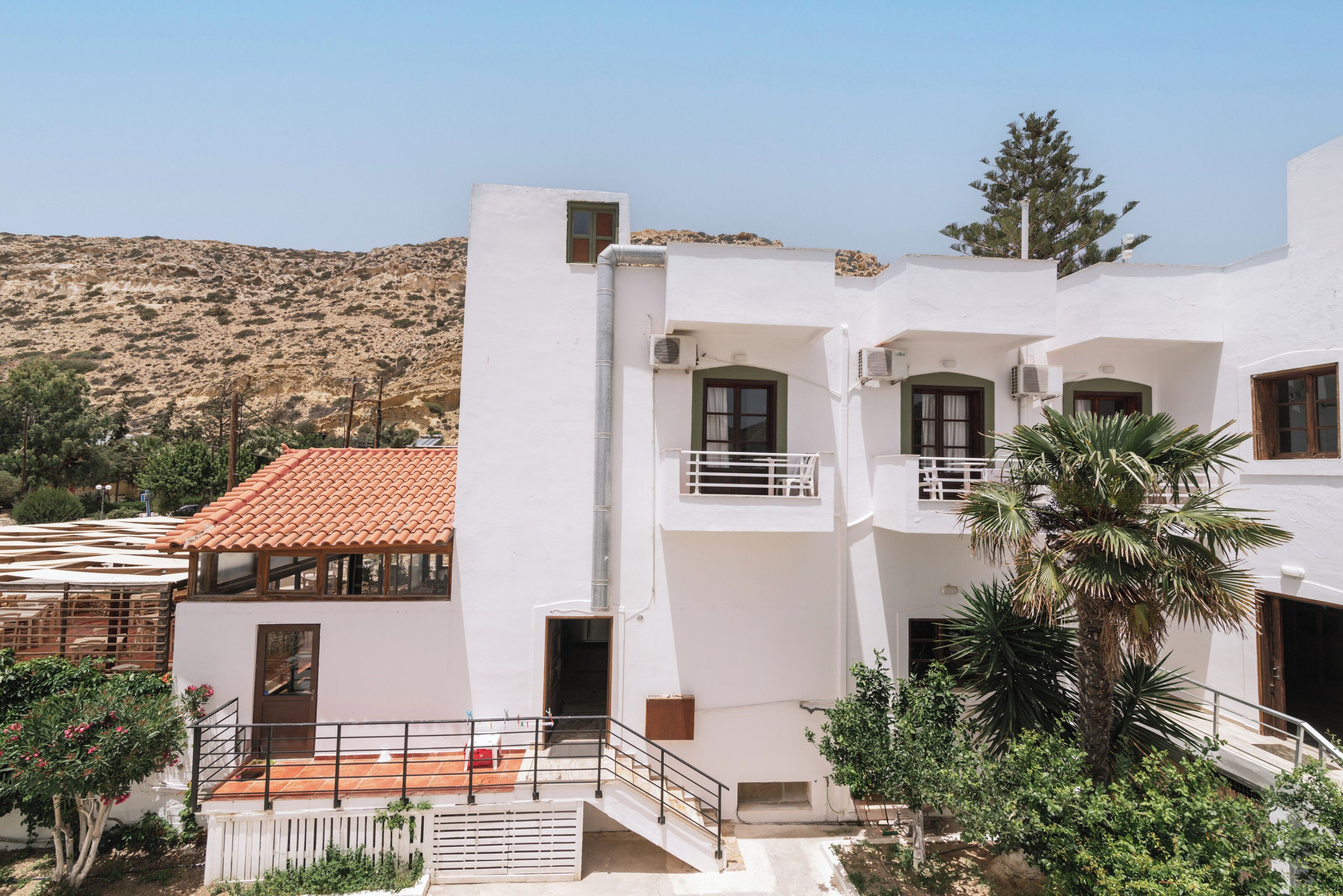 A white building with balconies, a red-tiled roof, and a mountainous backdrop.