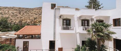 A white building with balconies, a red-tiled roof, and a mountainous backdrop.