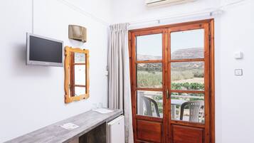 A room with a TV, a wooden-framed mirror, a small fridge, a window with curtains, and a view of a landscape.