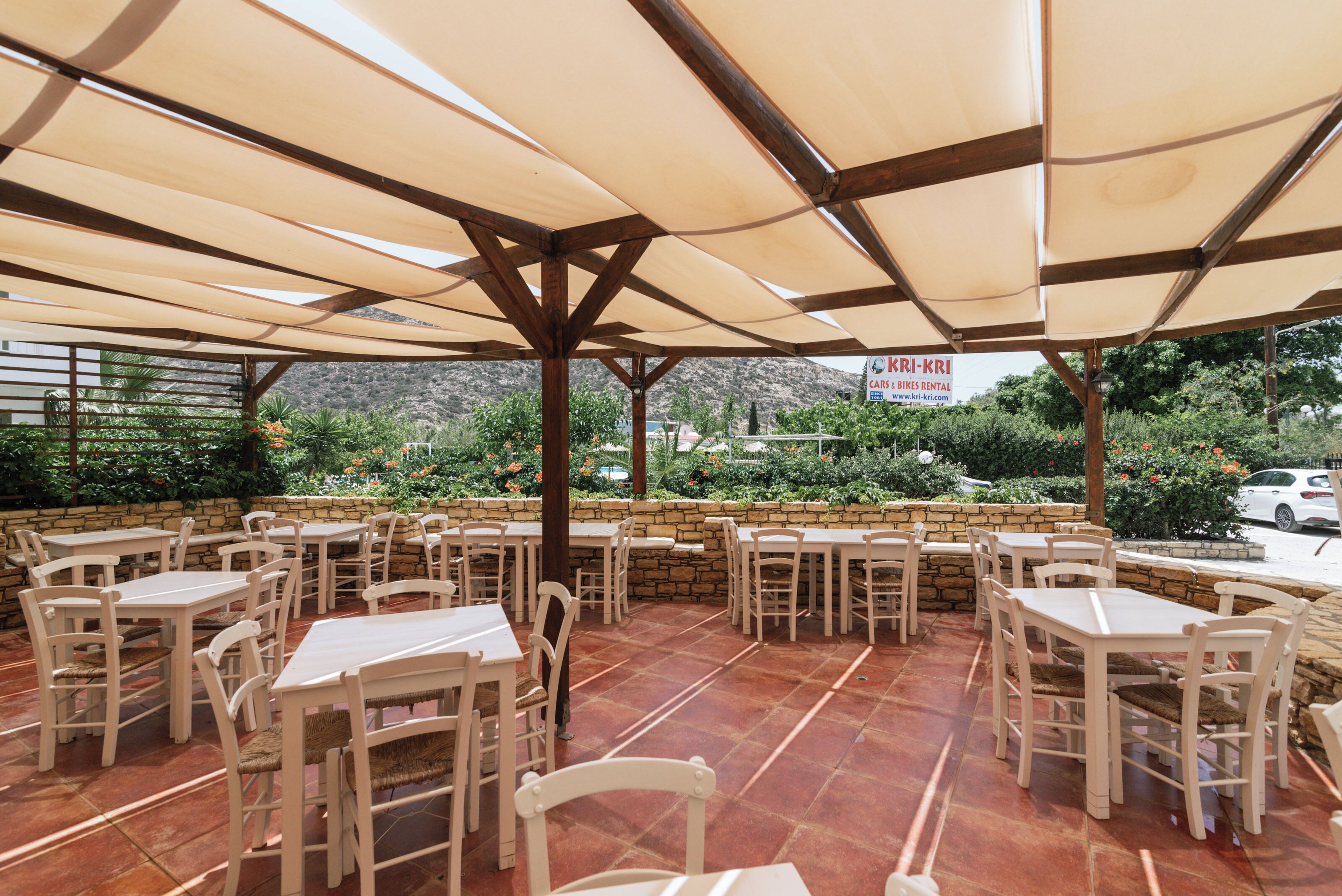 An outdoor patio with multiple tables and chairs, a stone wall, and a sign for KRI-KRI cars and bikes rental.