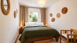 Een slaapkamer met een bed, bedlampen, een houten stoel, een rieten mand en een raam met gordijnen.