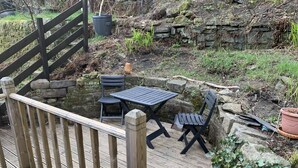 A wooden deck with a table and two chairs, surrounded by stone walls and greenery. - Meg's Den (Hebden Bridge)