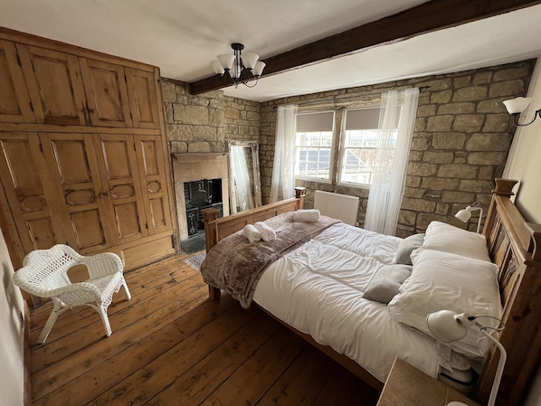 A bedroom with a stone wall, wooden floor, a bed with white linens, a wooden wardrobe, a white chair, and a fireplace.