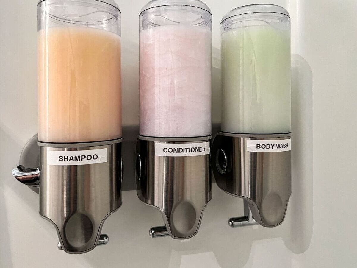Three stainless steel dispensers labeled 'SHAMPOO', 'CONDITIONER', and 'BODY WASH' mounted on a wall.