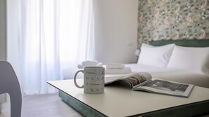 A hotel room with a bed, a bedside table with a magazine and a mug, and a window with curtains.