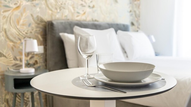 A neatly arranged dining table with a bowl and wine glass, set against a patterned wallpaper and a bed with white linens. - Italianway - The B Village Superior - 44 (Milan)