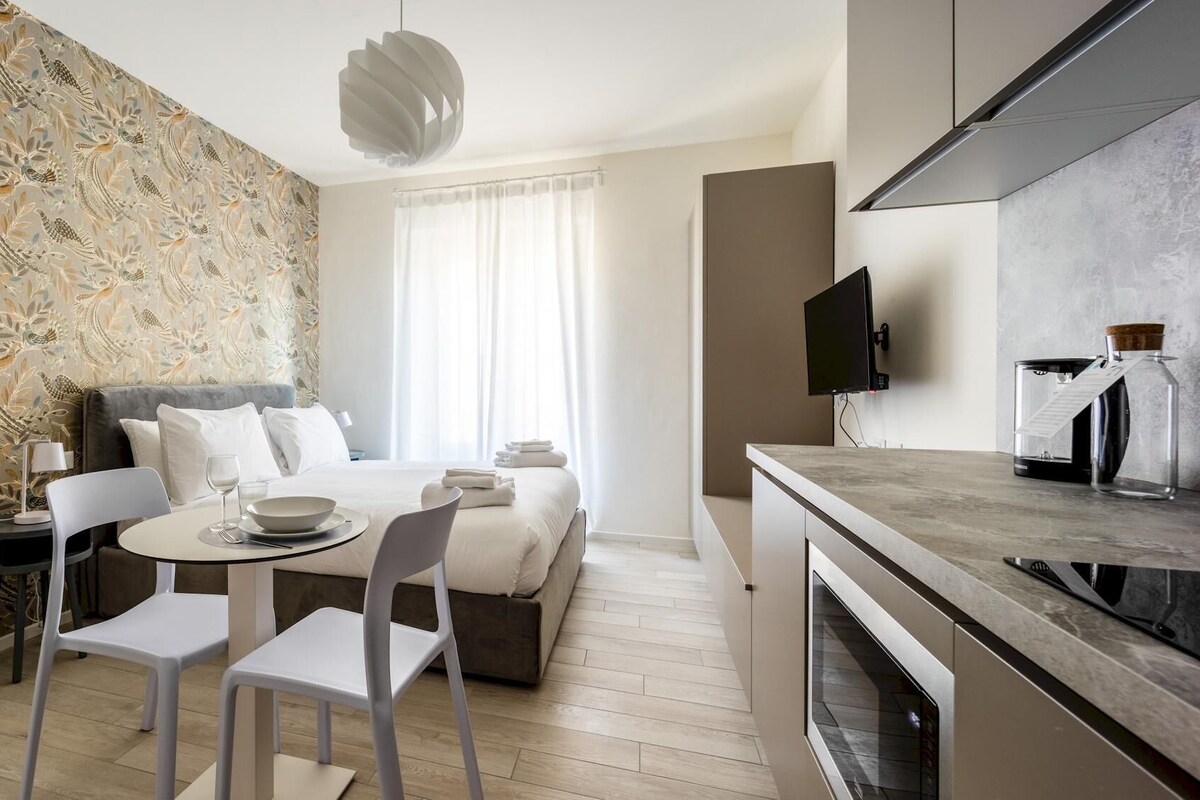 A modern hotel room with a bed, a dining area, a kitchen, and a large window with curtains.