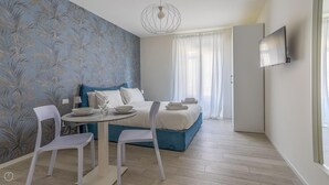 A modern bedroom with a bed, two white chairs, a small round table, and a TV mounted on the wall. - Italianway - The B Village Superior - 46 (Milan)