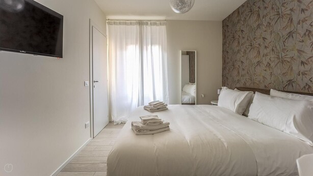 A hotel room with a large bed, a flat-screen TV mounted on the wall, a bathroom with a mirror, and a patterned wallpaper on the wall. - Italianway - The B Village Superior - 46 (Milan)
