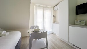 A hotel room with a bed, a dining table with a bowl and wine glass, a chair, a TV on the wall, and a window with curtains. - Italianway - The B Village Superior - 50 (Milan)