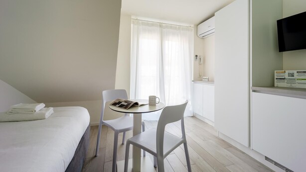 A hotel room with a bed, a small table with chairs, a window with curtains, an air conditioning unit, and a mounted TV. - Italianway - The B Village Superior - 50 (Milan)