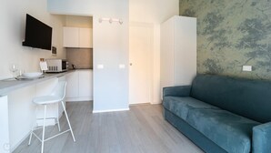 Een moderne keuken met witte kasten, een magnetron en een barkruk. Er staat een turquoise bank en een deur naar een andere kamer.