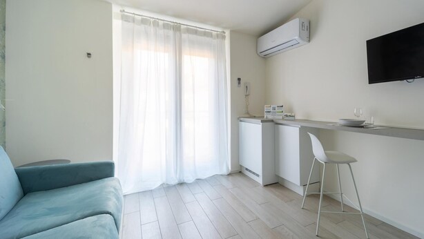 Een moderne, minimalistische woonkamer met een lichtblauwe bank, een keuken met een wit aanrecht en een wandgemonteerde flatscreen-televisie.