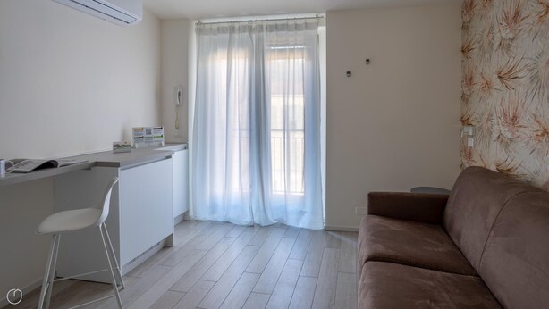 A small, bright room with a kitchenette, a white chair, a brown sofa, and a window with sheer curtains.