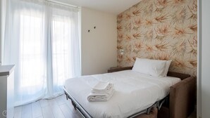A hotel room with a bed, a bedside table, a window with sheer curtains, and a floral wallpaper on the wall.