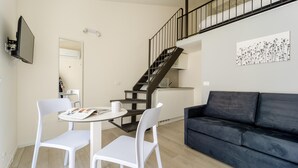 Un salon moderne avec un canapé foncé, des chaises blanches, une petite table sur laquelle se trouvent un magazine et une tasse, un téléviseur fixé au mur, un escalier et un coin cuisine.