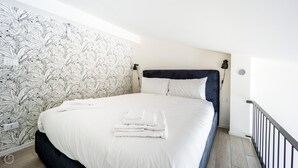 Un lit bien fait, avec du linge de lit blanc et des serviettes pliées, une tête de lit sombre et un mur orné d’un papier peint à motifs végétaux.