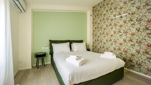 Una cama bien hecha con sábanas blancas y dos almohadas, una mesita de noche pequeña y una pared con estampado floral.