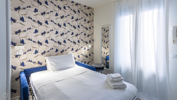A hotel room with a bed, a bedside table, a mirror, and a window with curtains. - Italianway - The B Village Business - A22 (Milan)
