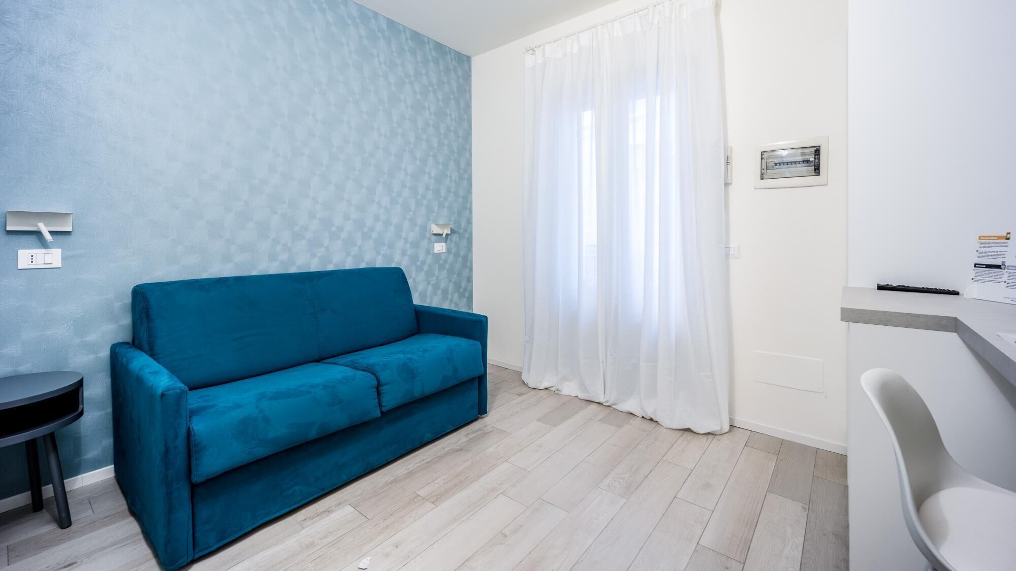 A room with a blue sofa, a small table, a white chair, and a window with white curtains.