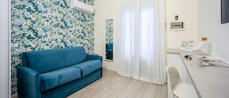 A room with a blue sofa, a white desk, and a large window with white curtains.