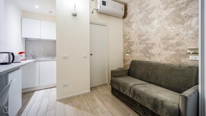 Un espace de vie compact comprenant un coin cuisine, un canapé gris et un mur orné d’un motif.