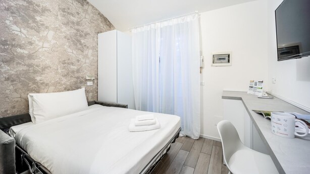 Ein Hotelzimmer mit einem Bett, einem Schreibtisch mit Stuhl, einem Fernseher und einem Fenster mit Vorhängen.
