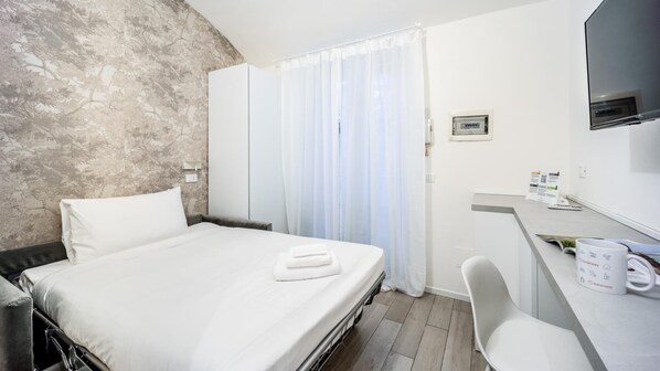 A hotel room with a bed, a desk with a chair, a TV, and a window with curtains. - Italianway - The B Village Standard - At03 (Milan)