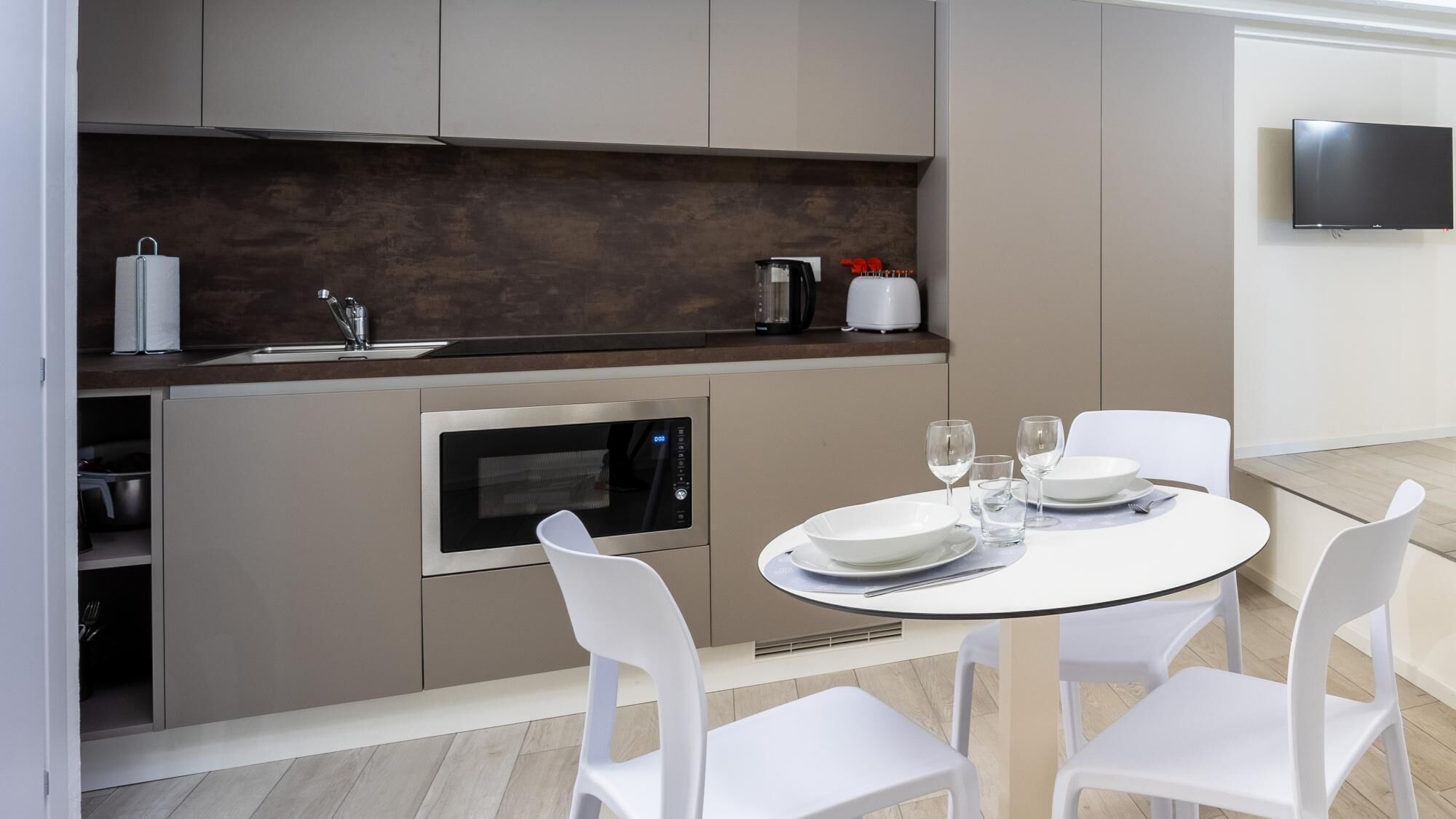 Une cuisine moderne équipée d’un four encastré, d’une machine à café et d’un évier. On y trouve une table à manger avec des chaises blanches et un téléviseur fixé au mur.